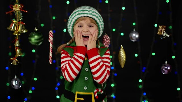 Overjoyed teen kid girl in Christmas elf Santa Claus helper costume laughs happily hears funny anecdote isolated on black background with garland. Child cannot stop laughing. Holidays celebration