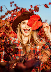 Beautiful young girl in a hat. Behind it is a background of red grape leaves. Autumn photo, a lot of emotions on her face, a grape leaf in her hand, she closes one eye with it and smiles beautifully.