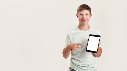 Happy disabled boy with Down syndrome smiling, holding and pointing at tablet pc with blank screen while standing isolated over white background