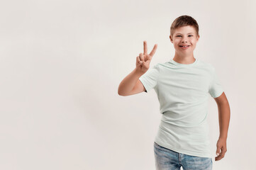Happy disabled boy with Down syndrome smiling at camera, showing peace sign with one hand while standing isolated over white background