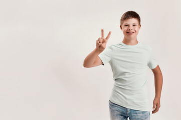 Happy disabled boy with Down syndrome smiling at camera, showing peace sign with one hand while...