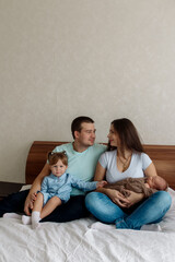 Young beautiful caucasian family with a three-year-old daughter and a newborn toddler sits embracing on the bed in the bedroom at home
