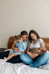 Young beautiful caucasian family with a three-year-old daughter and a newborn toddler sits embracing on the bed in the bedroom at home
