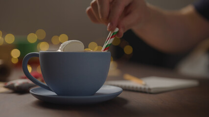 man put marshmallow in hot chocolate in blue cup in the evening with shopping list for holidays in notepad