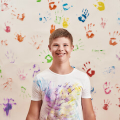Obraz na płótnie Canvas Cheerful disabled boy with Down syndrome smiling at camera while standing in front of the wall with many colorful hand prints