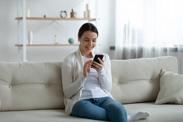 Smiling young woman using smartphone, sitting on couch at home, looking at screen, happy beautiful girl chatting with friends in social network, enjoying leisure time with phone, shopping online