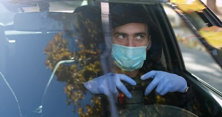 A driver in a face mask and blue gloves hugs the steering wheel of his car and looks at the working windshield washers, with yellow leaves. - Powered by Adobe