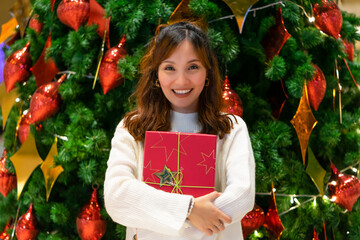 Young Asian woman in white sweater hugging red present box with a smiling face and happiness during the season in front of Christmas tree with glitter decoration. Merry Christmas and Happy Holidays.