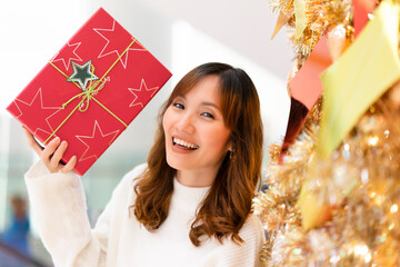Young woman holding red present box in hand, standing at Christmas Tree with glitter decoration, feeling happy and cheerful celebration of Christmas season. Merry Christmas and Happy Holidays.