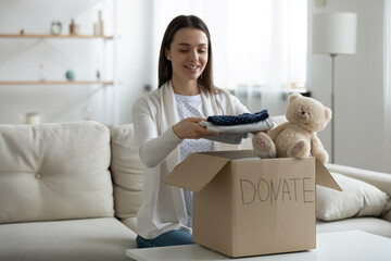 Smiling young woman putting clothing into donation box, sitting on couch at home, happy candid girl packing toys and clothes for children, helping to poor people, charity and aid concept