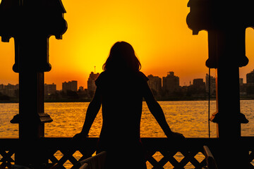 A young woman looking at the orange sunset on the Nile river, the sun is hiding in the buildings of...