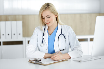 Woman-doctor at work while sitting at the desk in hospital or clinic. Blonde cheerful physician filling up medication history record form