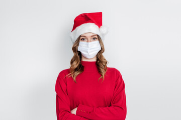 Fototapeta na wymiar A serious girl in a New Year's cap, a red sweater wearing a medical mask stands isolated on a white background. 2021 new year celebration concept