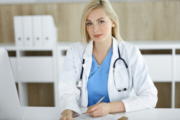 Portrate of woman-doctor at work while sitting at the desk in clinic. Blonde cheerful physician filling up medical form or prescription