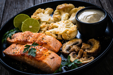 Roasted salmon steaks with fried cauliflower, fried brown champignons, lime and mayonnaise on wooden table
