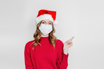 Fototapeta na wymiar Caucasian girl in santa hat, red sweater wearing medical protective face mask points finger at copy of text isolated on white studio background. Coronavirus quarantine concept