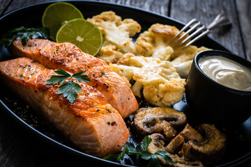 Roasted salmon steaks with fried cauliflower, fried brown champignons, lime and mayonnaise on wooden table
