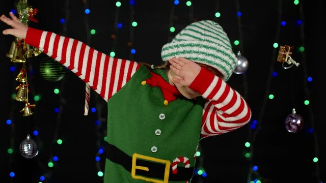 Teen kid girl in Christmas elf Santa Claus helper costume dancing and showing DAB dance gesture isolated on black background with garland. Child internet meme dancer. New Year holidays celebration