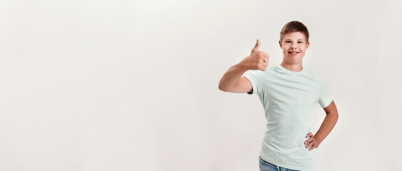Joyful disabled boy with Down syndrome smiling at camera, showing thumbs up with one hand while standing isolated over white background