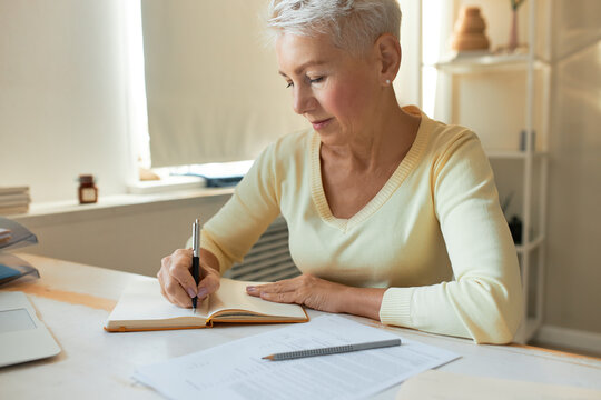 Attractive short haired mature female coach sitting at her workplace, writing in diary, making list, having concentrated look. Middle aged businesswoman noting down in copybook, working in office