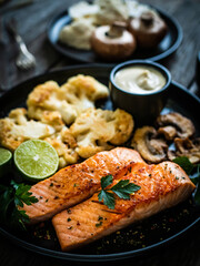 Roasted salmon steaks with fried cauliflower, fried brown champignons, lime and mayonnaise on wooden table
