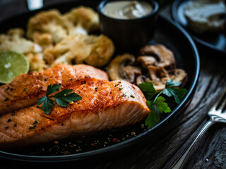 Roasted salmon steaks with fried cauliflower, fried brown champignons, lime and mayonnaise on wooden table
