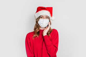Fototapeta na wymiar Cute caucasian student girl wearing a medical mask and santa hat red sweater holds hand near face isolated on white studio background