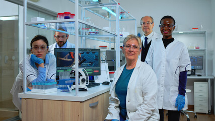 Team of multiethnic scientists sitting in laboratory looking at camera in modern equipped lab. Group of doctors examining virus evolution using high tech for scientific research, vaccine development.