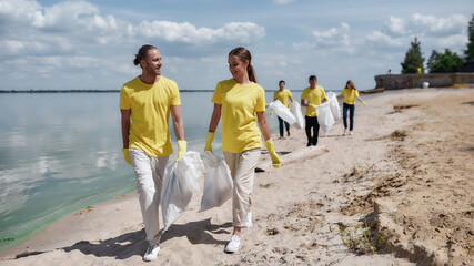 Two young happy friends eco activists wearing uniform and rubber gloves carrying trash bag while cleaning the beach with group of volunteers