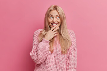 Joyful blonde woman with thoughtful happy expression concentrated above smiles broadly shows white teeth wears warm winter sweater isolated over pink background. Positive human emotions concept.