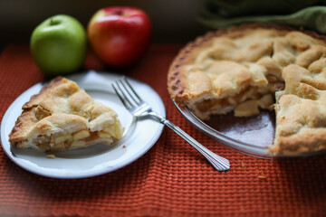 Slice of apple pie on tale with whole pie and apples.