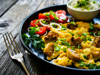 Delicious breakfast - scrambled eggs with fried champignon, cottage cheese and vegetable served on black plate on wooden table
