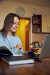 Young cheerful confident and happy freelancer woman is working on laptop at her kitchen at home