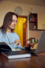 Young cheerful confident and happy freelancer woman is working on laptop at her kitchen at home