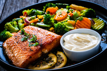Fried salmon fillet with mix of vegetables, lemon and mayonnaise served on black plate on wooden table
