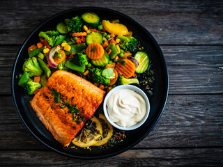 Fried salmon fillet with mix of vegetables, lemon and mayonnaise served on black plate on wooden table
