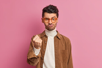 Handsome adult man clenches fist and expresses negative emotions being annoyed with someone wears transparent glasses and fashionable brown shirt isolated over pink background. I will show you