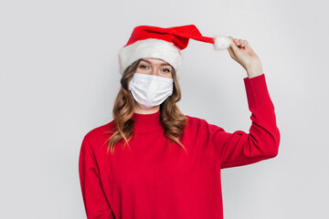 Fototapeta na wymiar Happy playful young woman in santa hat, red sweater and medical protective respiratory face mask holding and pulling balabon isolated on gray studio background