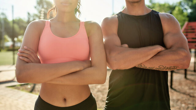 Cropped shot of sportive young couple, man and woman standing with arms crossed while having workout together at outdoor gym