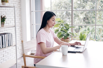 Asian businesswoman in white shirt is smiling and online working with laptop at home. She is blogger freelance in social media website. Communication and technology concept.