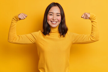 Pretty young brunette Asain woman raises arms and shows biceps demonstrates muscles feels proud about personal achievements wears yellow turtleneck stands indoor. People strength and power concept