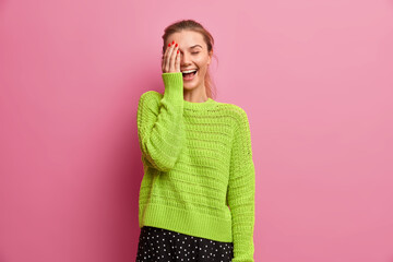 Studio shot of happy millennial girl makes face palm and laughs joyfully wears knitted jumper isolated over pink background. Beautiful European woman with pony tail expresses happiness and joy