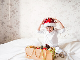 Fototapeta na wymiar Joyful boy in Santa Claus hat is playing with decorations for Christmas tree. Funny kid is ready for New Year celebration. Cozy home. Winter holiday spirit.