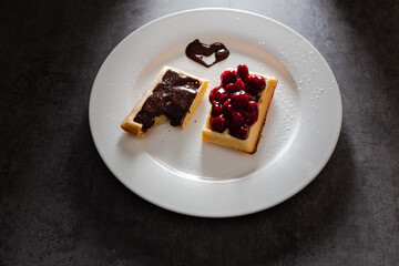 Tasty waffles with fruit and chocolate topping, white plate on dark background under directional back light.