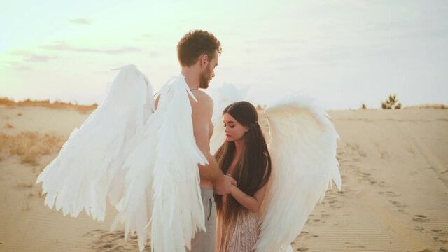 embrace hug heaven angels with white wings. man holding a woman's hand. girl looks at guy with love in her eyes, innocent beautiful face. Model fantastic costume. Backdrop bright sunny sunset desert