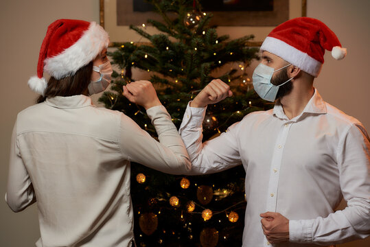 Elbow Greeting To Avoid The Spread Of Covid-19 Against The Background Of A Christmas Tree At Home. A Man And A Woman In Medical Masks And Santa Hats. Instead Of A Hug Or A Handshake, They Bump Elbows.