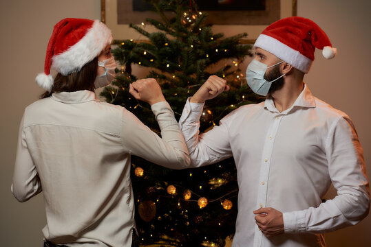 Elbow Greeting To Avoid The Spread Of Coronavirus Against The Background Of A Christmas Tree At Home. A Man And A Woman In Medical Masks And Santa Hats. Instead Of A Hug Or Handshake, They Bump Elbows