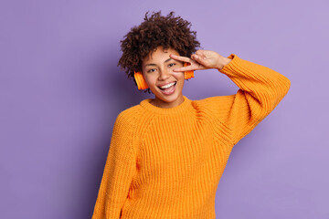 Obraz na płótnie Canvas Joyful relaxed dark skinned millennial girl makes victory gesture listens favorite music in wireless headphones wears knitted orange jumper isolated over purple background chooses song to listen