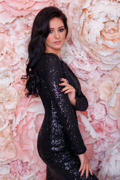 Young woman with long curly brunette hair in black festive dress on flower wall background