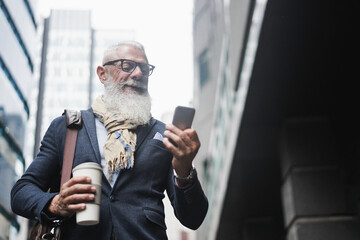 Business hipster senior man using mobile phone and drinking coffee with city in background - Focus...
